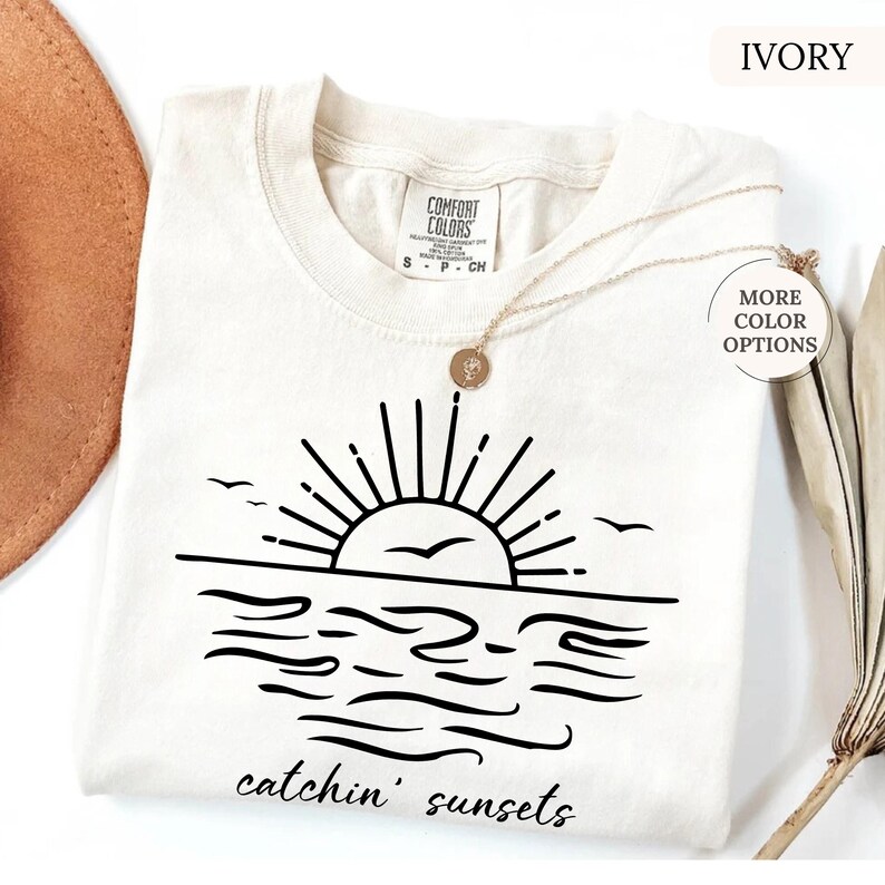 May include: Ivory-colored t-shirt with a black graphic of a sunset over the ocean, featuring the text "catchin' sunsets." The shirt has a crew neck and short sleeves. A gold necklace with a pendant is draped on the shirt.