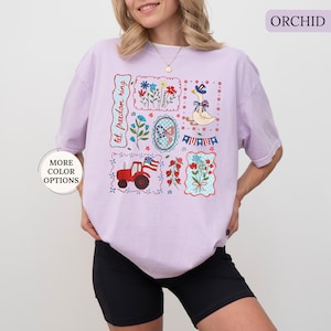 May include: Orchid-colored t-shirt featuring a patchwork design with patriotic and floral illustrations. The design includes a red tractor, an American flag, a goose wearing a hat, and the text "let freedom ring."