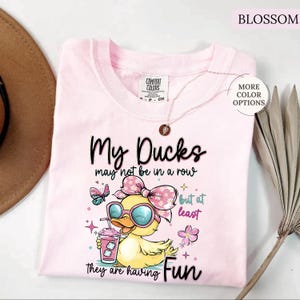 May include: Light pink t-shirt with the text "My Ducks may not be in a row but at least they are having Fun." The shirt features a cartoon duck wearing sunglasses and a bow, holding a drink. The word "Blossom" is at the top.