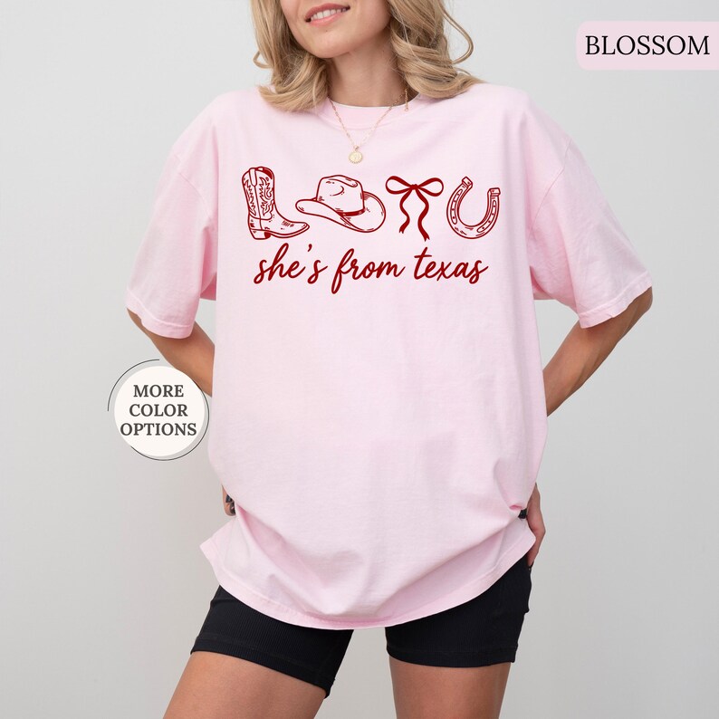 Pode incluir: Camiseta rosa claro com um gr&aacute;fico vermelho de bota de cowboy, chap&eacute;u, la&ccedil;o e ferradura, com o texto "she's from texas". A camiseta tem um corte folgado e &eacute; de cor lisa. As palavras "More Color Options" est&atilde;o em um c&iacute;rculo.