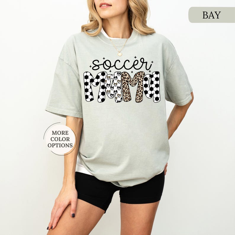 Pu&ograve; includere: T-shirt verde chiaro con la scritta "soccer mama" in un font giocoso. La parola "soccer" &egrave; in nero con motivi a pallone da calcio bianchi, e "mama" ha un mix di motivi, tra cui stampa leopardata.