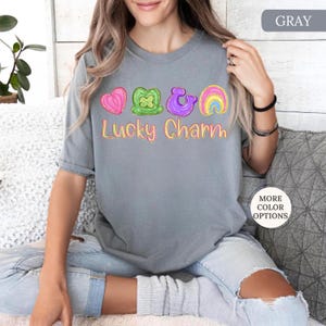 May include: Gray t-shirt with the text "Lucky Charm" and colorful graphic icons. The icons include a pink heart, green shamrock, purple horseshoe, and a rainbow. The shirt is a casual style.