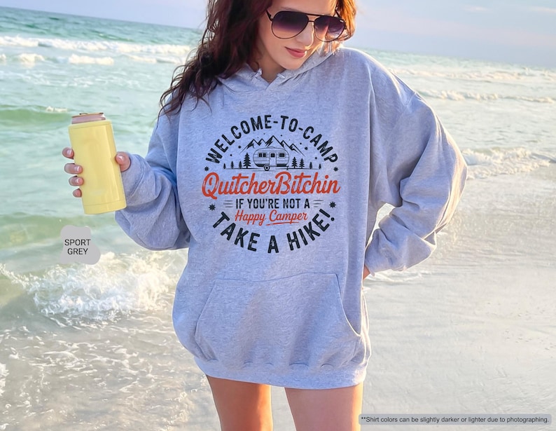 May include: A light grey hoodie with a front pocket and the text "WELCOME-TO-CAMP Quitcher Bitchin IF YOU'RE NOT A Happy Camper TAKE A HIKE!". The hoodie is paired with a yellow can cooler. The words "SPORT GREY" are also visible.