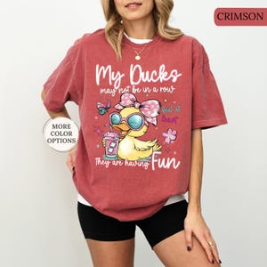 May include: Crimson-colored t-shirt with the text "My Ducks may not be in a row but at least they are having Fun." The graphic features a cartoon duck wearing sunglasses and a bow, holding a drink. Additional color options are available.