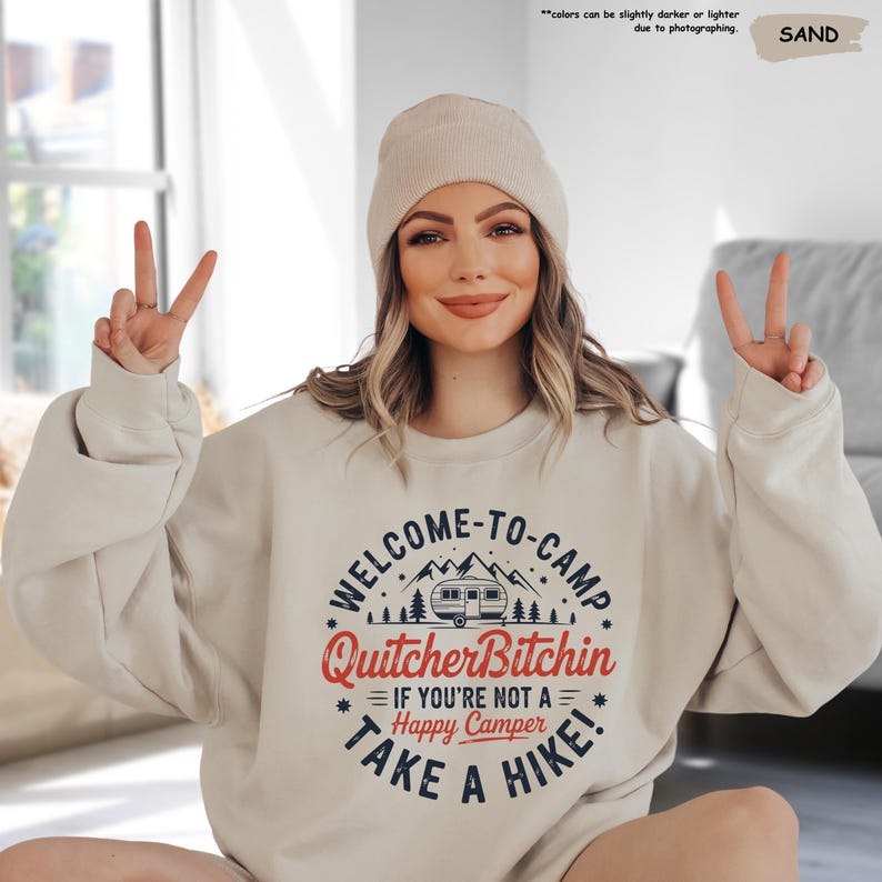 May include: Sand-colored sweatshirt with the text "WELCOME-TO-CAMP Quitcher Bitchin IF YOU'RE NOT A Happy Camper TAKE A HIKE!" in a circular design. The design includes a camper, mountains, and trees.