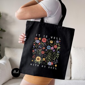 May include: Black tote bag with a floral design and the text "IT IS WELL WITH MY SOUL" in white. The floral design features colorful flowers and foliage. The bag has black handles and a black base.