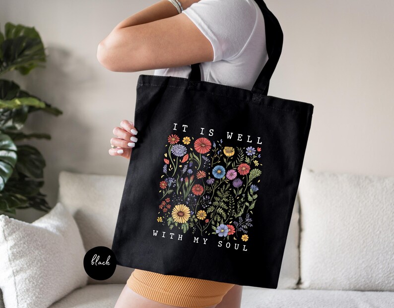 May include: Black tote bag with a floral design and the text "IT IS WELL WITH MY SOUL" in white. The floral design features colorful flowers and foliage. The bag has black handles and a black base.
