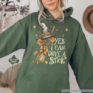 Op de afbeelding: Militairgroene hoodie met een heksenhoed, bezem en herfstbladeren. De tekst "YES I CAN DRIVE A STICK" is op de voorkant gedrukt. De hoodie heeft een capuchon met trekkoord en een voorzak.