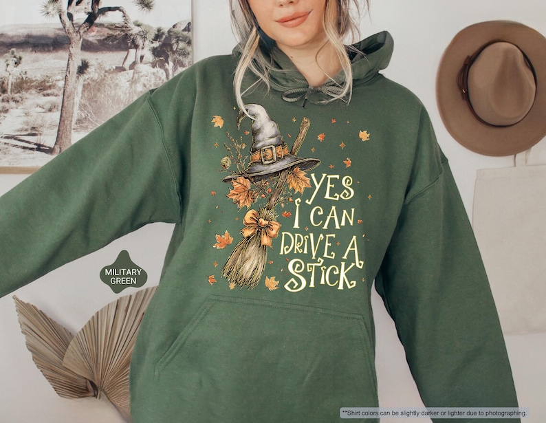 Op de afbeelding: Militairgroene hoodie met een heksenhoed, bezem en herfstbladeren. De tekst "YES I CAN DRIVE A STICK" is op de voorkant gedrukt. De hoodie heeft een capuchon met trekkoord en een voorzak.
