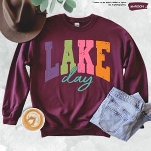 Op de afbeelding: Een bordeauxrode sweatshirt met de tekst "LAKE day" in kleurrijke, versleten letters. Het woord "LAKE" is in paars, groen, roze en oranje. Het woord "day" is turquoise. De sweatshirt wordt gecombineerd met een blauwe jeans en een bruine hoed.