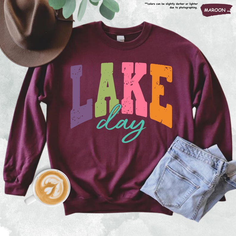 Op de afbeelding: Een bordeauxrode sweatshirt met de tekst "LAKE day" in kleurrijke, versleten letters. Het woord "LAKE" is in paars, groen, roze en oranje. Het woord "day" is turquoise. De sweatshirt wordt gecombineerd met een blauwe jeans en een bruine hoed.