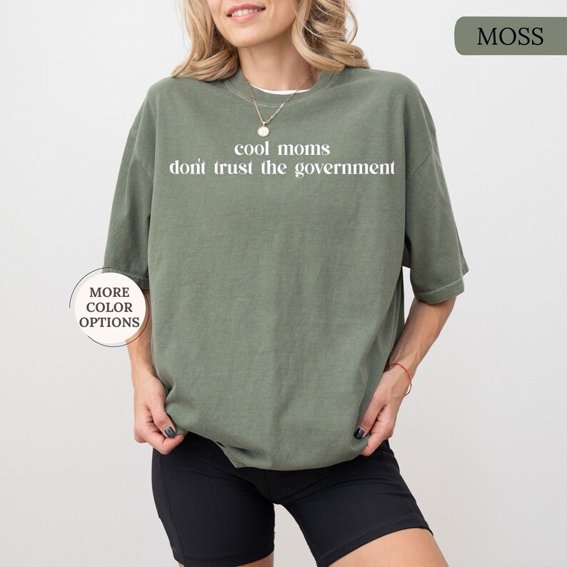 Peut inclure: T-shirt vert mousse avec l'inscription blanche "cool moms don't trust the government". Le t-shirt est ample et &agrave; manches courtes. Le mod&egrave;le porte un short noir. Un bouton sur le t-shirt indique "More Color Options".