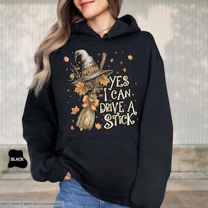 Op de afbeelding: Zwarte hoodie met een afbeelding van een heksenhoed, bezem en de tekst "YES I CAN DRIVE A STICK". Het ontwerp bevat herfstbladeren en een strik. De hoodie heeft een voorzak.