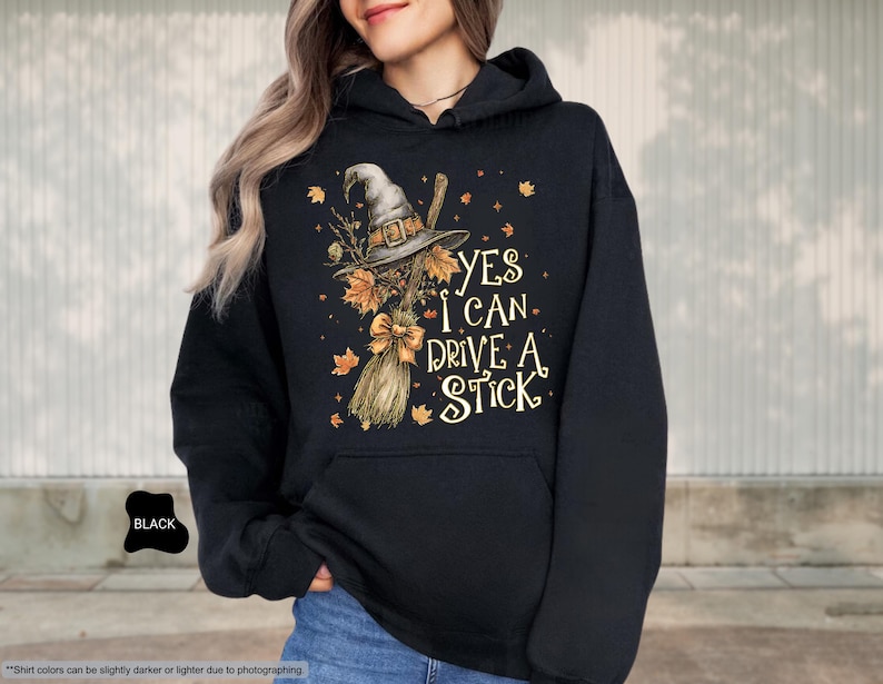 Op de afbeelding: Zwarte hoodie met een afbeelding van een heksenhoed, bezem en de tekst "YES I CAN DRIVE A STICK". Het ontwerp bevat herfstbladeren en een strik. De hoodie heeft een voorzak.