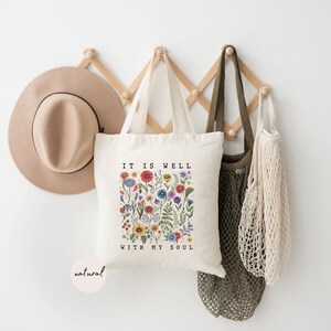 May include: A beige tote bag with the text "IT IS WELL WITH MY SOUL" and a floral design. A tan hat and several bags hang on a wooden rack against a white wall. The tote bag has a natural color.