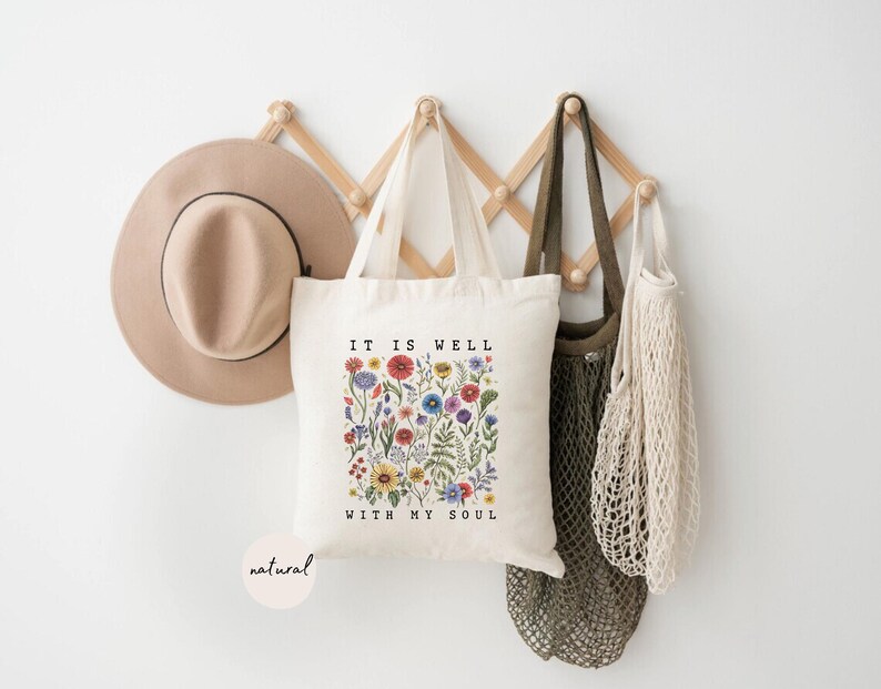 May include: A beige tote bag with the text "IT IS WELL WITH MY SOUL" and a floral design. A tan hat and several bags hang on a wooden rack against a white wall. The tote bag has a natural color.