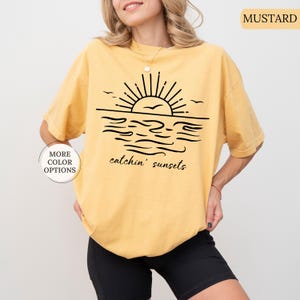 May include: Mustard-colored t-shirt featuring a black graphic of a sunset over the ocean with the text "catchin' sunsets." The shirt has short sleeves and a relaxed fit. Additional color options are available.