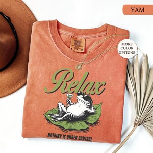 May include: A yam-colored t-shirt with a graphic of a frog wearing sunglasses, holding a cocktail, and lounging on a lily pad. The word "Relax" is in green script above the frog, and the phrase "Nothing is Under Control" is below.