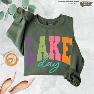 Op de afbeelding: Militairgroen sweatshirt met de tekst "LAKE day" in kleurrijke blokletters. Het woord "LAKE" is paars, groen, roze en oranje. Het woord "day" is turquoise. De sweatshirt is opgevouwen en heeft een ronde hals.