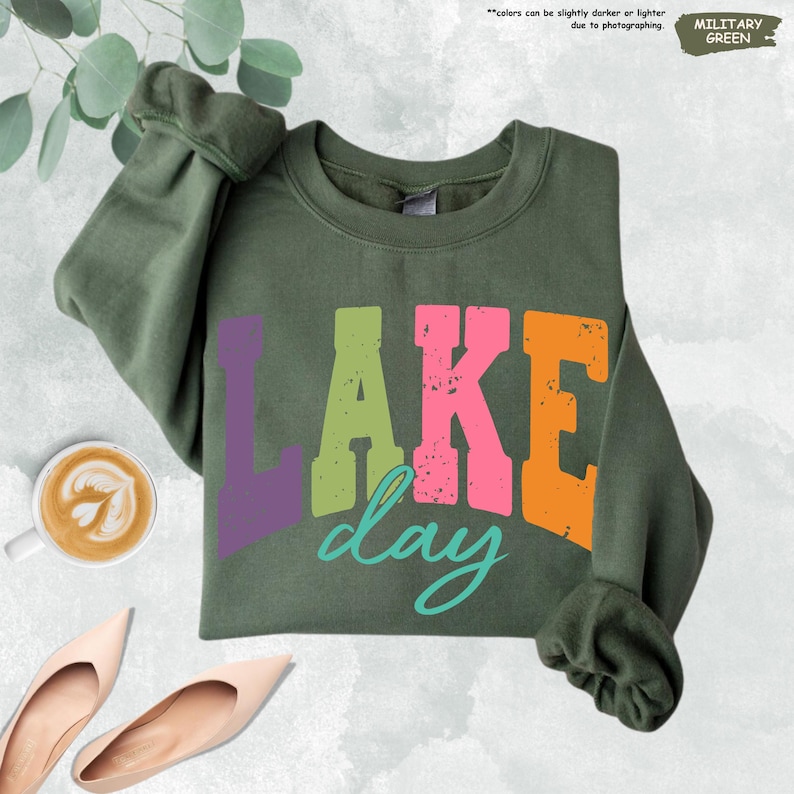 Op de afbeelding: Militairgroen sweatshirt met de tekst "LAKE day" in kleurrijke blokletters. Het woord "LAKE" is paars, groen, roze en oranje. Het woord "day" is turquoise. De sweatshirt is opgevouwen en heeft een ronde hals.