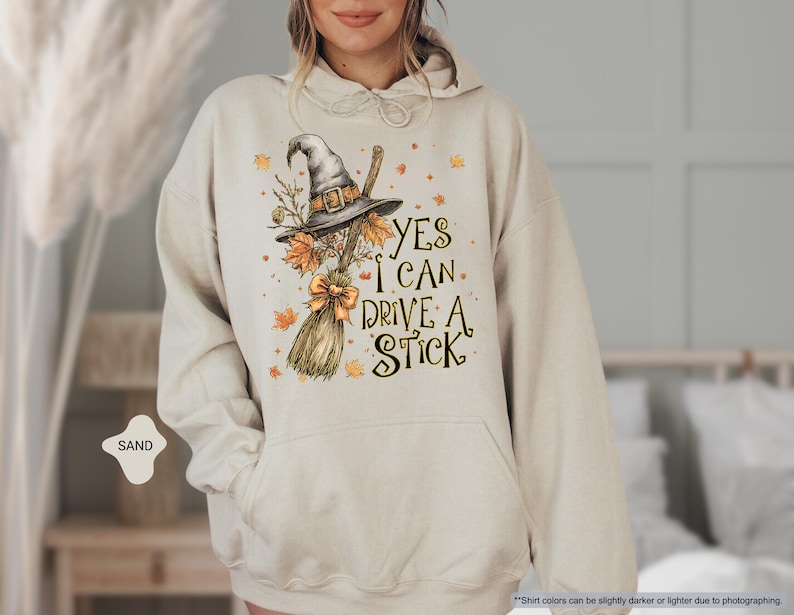 Op de afbeelding: Zandkleurige hoodie met een heksenhoed, bezem en herfstblad-afbeelding. De tekst luidt "YES I CAN DRIVE A STICK". Het ontwerp bevat een strik en decoratieve elementen.