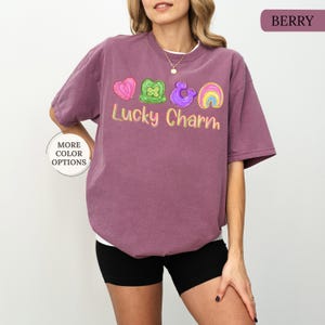 May include: Berry-colored t-shirt with the text "Lucky Charm" and colorful graphics of a heart, shamrock, horseshoe, and rainbow. The shirt has a crew neck and short sleeves. The words "More Color Options" are in a circle on the left side.