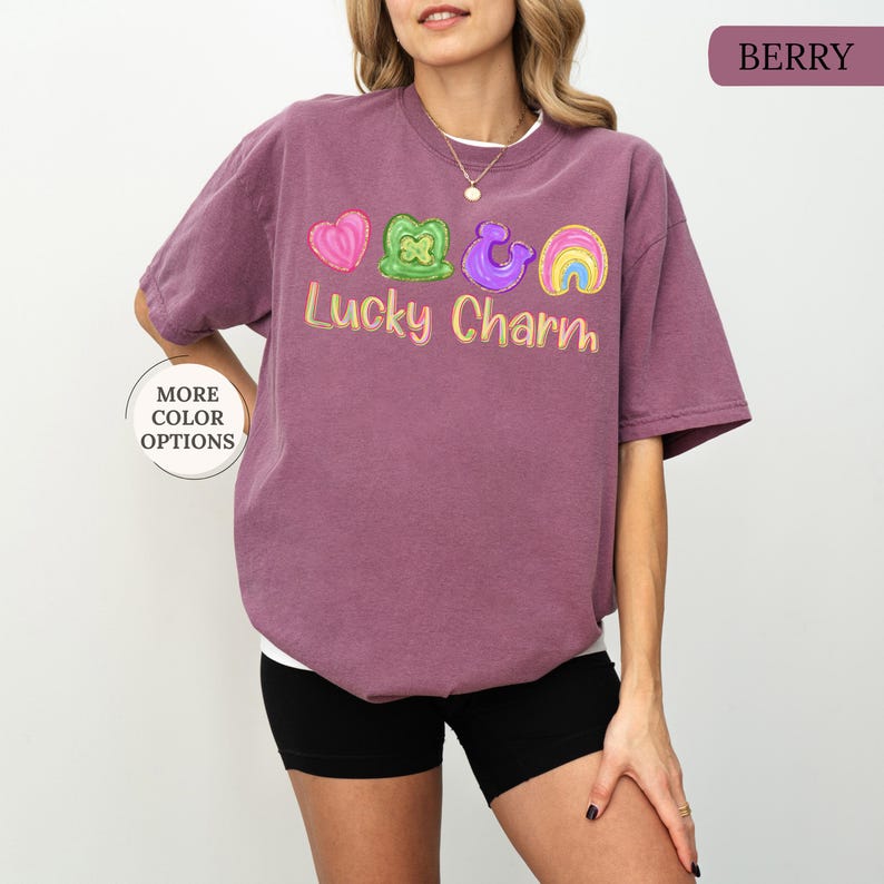 May include: Berry-colored t-shirt with the text "Lucky Charm" and colorful graphics of a heart, shamrock, horseshoe, and rainbow. The shirt has a crew neck and short sleeves. The words "More Color Options" are in a circle on the left side.