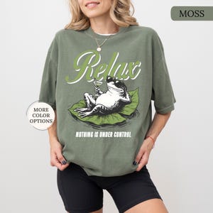 May include: Moss green t-shirt with a graphic of a frog wearing sunglasses, relaxing on a lily pad and holding a cocktail. The word "Relax" is above the frog, and "Nothing is Under Control" is below.