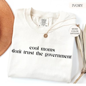 Peut inclure: T-shirt ivoire avec l'inscription noire "cool moms don't trust the government". Le t-shirt a un col rond et des manches courtes. Un collier dor&eacute; avec un pendentif est drap&eacute; sur le t-shirt.
