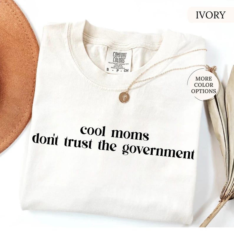 Peut inclure: T-shirt ivoire avec l'inscription noire "cool moms don't trust the government". Le t-shirt a un col rond et des manches courtes. Un collier dor&eacute; avec un pendentif est drap&eacute; sur le t-shirt.