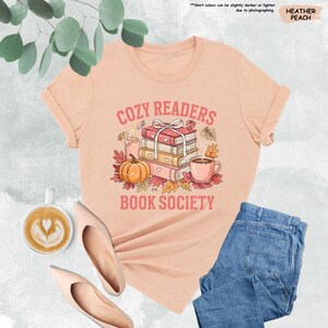May include: Heather peach-colored t-shirt with the text "COZY READERS BOOK SOCIETY" in a vintage style. The graphic includes a stack of books, a pumpkin, a candle, a coffee cup, and autumn leaves. The shirt is paired with blue jeans and beige shoes.