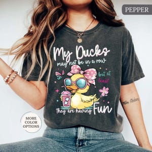 May include: Dark gray t-shirt with the text "My Ducks may not be in a row but at least they are having Fun." The shirt features a cartoon duck wearing sunglasses and a bow, holding a drink. Additional graphics include butterflies and flowers.