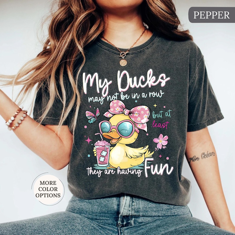 May include: Dark gray t-shirt with the text "My Ducks may not be in a row but at least they are having Fun." The shirt features a cartoon duck wearing sunglasses and a bow, holding a drink. Additional graphics include butterflies and flowers.