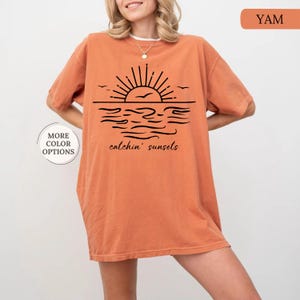 May include: Orange-colored t-shirt with a black graphic of a sunset over water, with the words "catchin' sunsets." The shirt has short sleeves and a relaxed fit. The text "MORE COLOR OPTIONS" is also visible.
