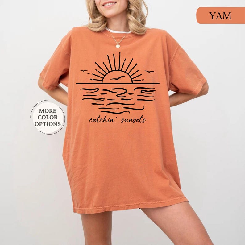 May include: Orange-colored t-shirt with a black graphic of a sunset over water, with the words "catchin' sunsets." The shirt has short sleeves and a relaxed fit. The text "MORE COLOR OPTIONS" is also visible.