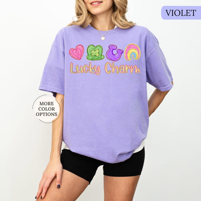 May include: A violet short-sleeved t-shirt with the text "Lucky Charm" and colorful graphic designs of a heart, shamrock, horseshoe, and rainbow. The shirt has a relaxed fit and is worn with black shorts.