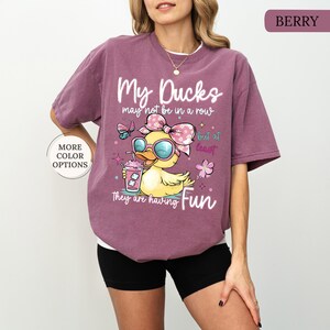 May include: Berry-colored t-shirt with a graphic of a cartoon duck wearing sunglasses and a bow, holding a drink. Text reads "My Ducks may not be in a row but at least they are having Fun." The shirt has a relaxed fit.
