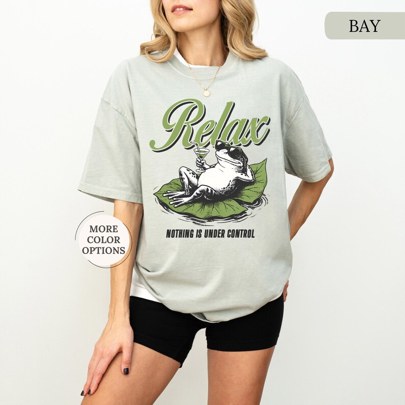 May include: Sage green t-shirt with a graphic of a frog relaxing on a lily pad, holding a cocktail. The word "Relax" is above the frog in green script, with the phrase "Nothing is Under Control" below.