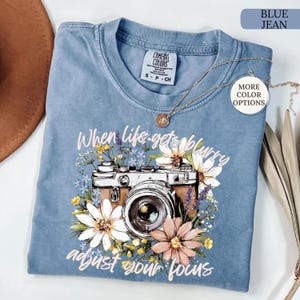 May include: Blue jean colored t-shirt with a floral camera graphic and the text "When life gets blurry, adjust your focus." The shirt has a crew neck and short sleeves. A gold necklace is draped across the shirt.