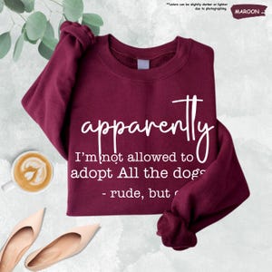May include: Maroon sweatshirt with the text "apparently I'm not allowed to adopt All the dogs - rude, but". The sweatshirt is a solid maroon color. A cup of coffee and a pair of shoes are in the image.