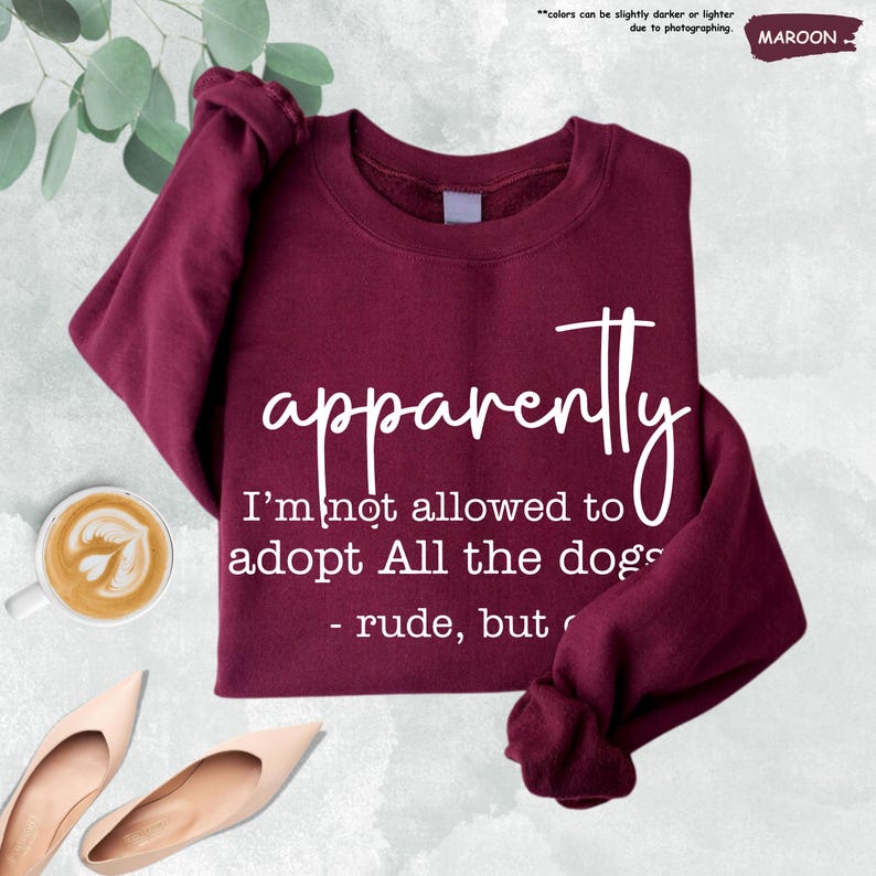 May include: Maroon sweatshirt with the text "apparently I'm not allowed to adopt All the dogs - rude, but". The sweatshirt is a solid maroon color. A cup of coffee and a pair of shoes are in the image.