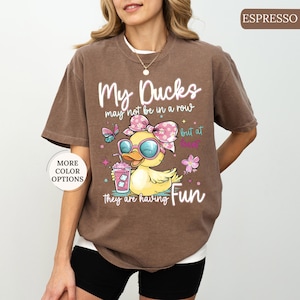 May include: Espresso-colored t-shirt with the text "My Ducks may not be in a row but at least they are having Fun." The graphic features a cartoon duck wearing sunglasses and a bow, holding a drink. Additional text says "More Color Options."