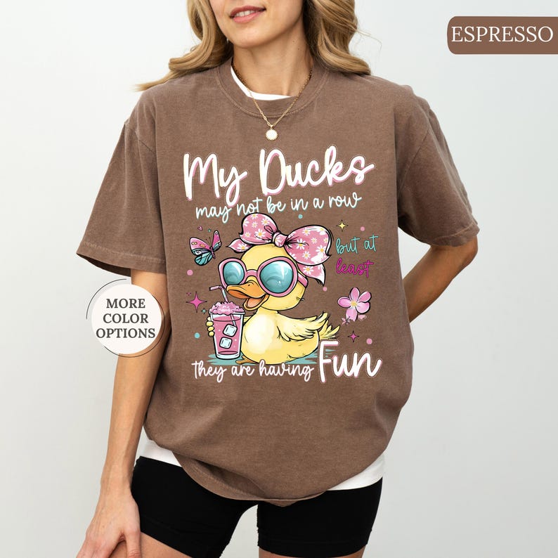 May include: Espresso-colored t-shirt with the text "My Ducks may not be in a row but at least they are having Fun." The graphic features a cartoon duck wearing sunglasses and a bow, holding a drink. Additional text says "More Color Options."