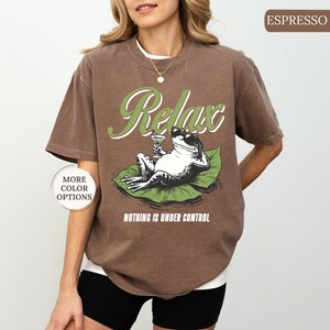 May include: Espresso-colored t-shirt featuring a relaxed frog graphic. The frog, wearing sunglasses and holding a cocktail, is lounging on a lily pad. The shirt has the word "Relax" in a green script and the phrase "Nothing is Under Control".