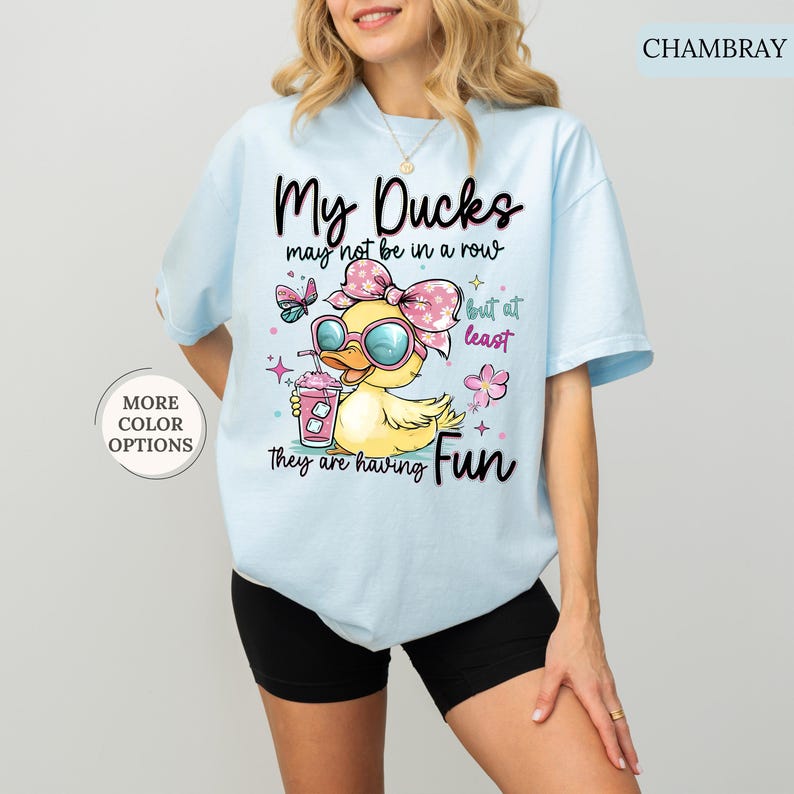 May include: Light blue t-shirt with a graphic of a yellow duck wearing sunglasses and a pink bow, holding a drink. Text reads "My Ducks may not be in a row but at least they are having Fun."