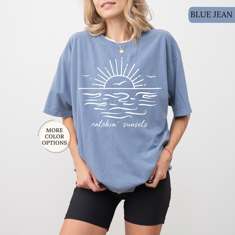 May include: Blue jean colored t-shirt with a white graphic of a sunset over the ocean, with the words "catchin' sunsets". The shirt has short sleeves and a relaxed fit. The words "MORE COLOR OPTIONS" are also visible.