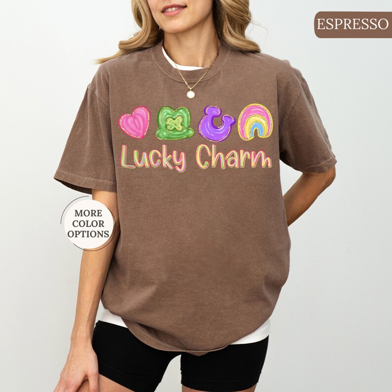 May include: Espresso-colored t-shirt with a white undershirt. The shirt features a graphic of a pink heart, green shamrock, purple horseshoe, and rainbow, with the words "Lucky Charm" in colorful letters. The shirt has short sleeves and a crew neck.
