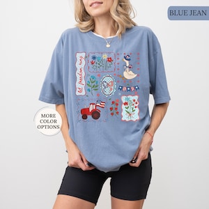 May include: Blue jean colored t-shirt with a white collar. The shirt features several embroidered patches with patriotic themes, including a tractor, flowers, a goose in a hat, and the text "let freedom ring."