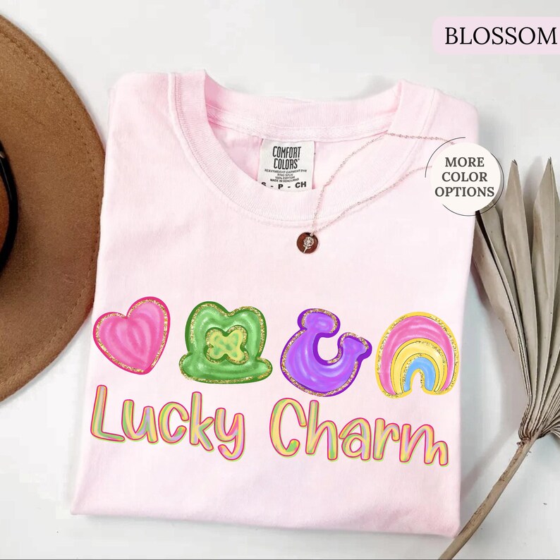 May include: A light pink t-shirt with the words "Lucky Charm" and colorful graphics of a heart, shamrock, ampersand, and rainbow. The shirt has a round neckline and short sleeves. A gold necklace with a pendant is visible.