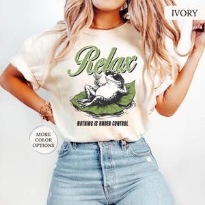 May include: Ivory-colored t-shirt featuring a relaxed frog graphic. The frog, wearing sunglasses and holding a cocktail, is lounging on a lily pad. The shirt has the word "Relax" above the frog and "Nothing is Under Control" below.